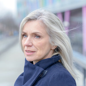 Ältere Frau mit grauem Haar und blauem Mantel blickt draußen nachdenklich zur Seite.